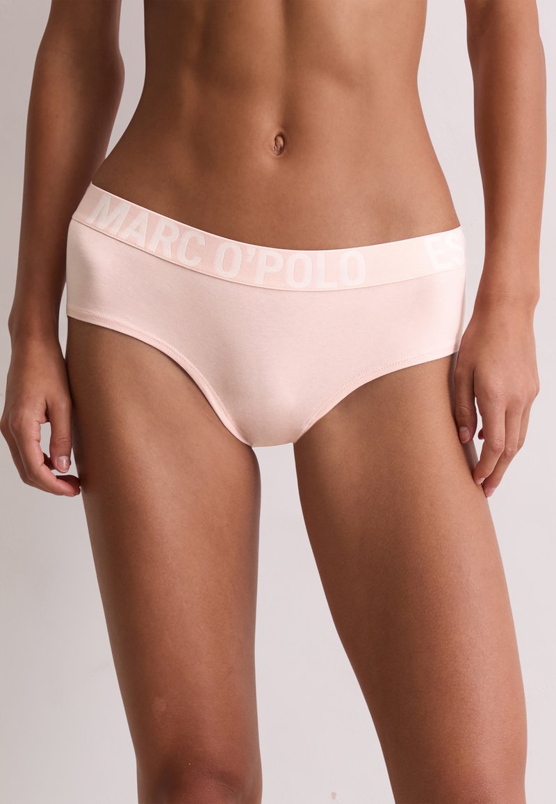 Kobieta nosząca jasnoróżową bieliznę w stylu hipster z elastycznym paskiem z dużymi literami z nazwą marki.