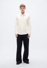 Μπλούζα εκρού με μακριά μανίκια και κουμπιά, εφαρμοστή γραμμή, συνδυασμένη με τζιν παντελόνι σε σκούρο ντένιμ με φαρδιά μπατζάκια και μαύρα παπούτσια. Μίνιμαλ σχεδιασμός, λεία υφή.