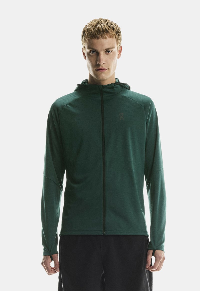 Joven vestido con una sudadera con capucha de color verde oscuro y cremallera, pantalones negros, de pie contra un fondo blanco liso.