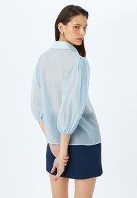 Blusa azul claro, transparente, con cuello tipo camisero, mangas abullonadas y diseño texturizado. Llevada sobre una mini falda ajustada de color oscuro.