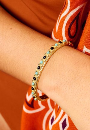 Bracelet en or avec des pierres turquoise et noires alternées, au design texturé et aux accents décoratifs, porté sur un poignet nu.