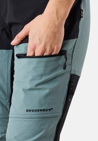 Vaaleansiniset ja mustat housut, joissa on vetoketjullinen sivutasku, varustettuna mustalla "SWEDEMOUNT"-logolla, valmistettu teksturoidusta materiaalista.