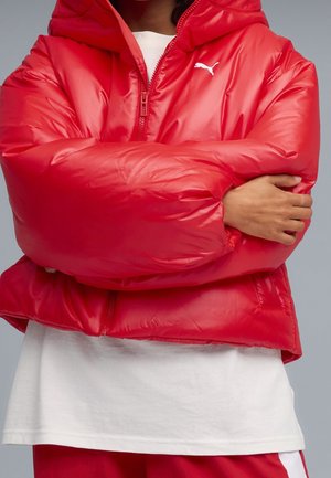 Person trägt eine glänzende rote Puma-Steppjacke mit verschränkten Armen über einem weißen Hemd und passenden roten Hosen.