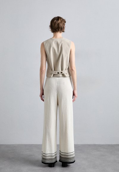 Beige mouwloze top met knoopdetail op de rug, gecombineerd met crèmekleurige wijde broek met textuurstreepjes en zwarte rand aan de onderkant.