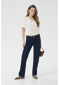 Blouse blanche avec des accents en dentelle, jean droit en denim foncé, ceinture marron et ballerines marron clair. Un petit sac à main marron est tenu.