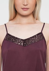 Burgundska bretela s tankim naramenicama, s dekolteom u obliku slova V ukrašenim šljokicama. Glatka tkanina s mekim teksturom, elegantan i prilagođen dizajn.