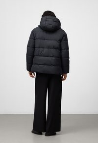 Giacca a vento nera con cappuccio, caratterizzata da un design trapuntato e una vestibilità ampia. Indossata sopra pantaloni neri a gamba larga e scarpe nere minimaliste.