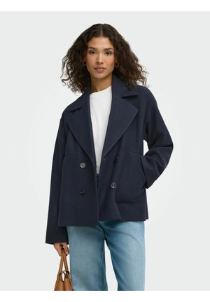 Femme aux cheveux bouclés portant un manteau bleu marine à double boutonnage, une chemise blanche, un jean bleu clair et tenant un sac à main marron dans sa main gauche.