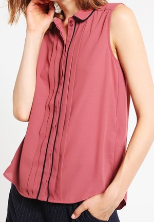 Ærmeløs rosa bluse med sort piping langs knapstolpe, båret af en person med den ene hånd, der rører ansigtet, og den anden i lommen.
