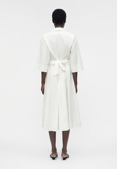 Άτομο που φοράει λευκό φόρεμα μήκους μέχρι το μέσο της γάμπας με τρία τέταρτα μανίκια και ζώνη δεμένη στη μέση, όρθιο ξυπόλητο σε απλό φόντο.