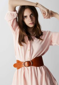 Helles rosa Oberteil mit Knöpfen, langen Ärmeln und lässigem Design, in der Taille mit einem breiten braunen Ledergürtel mit einer silbernen Schnalle gerafft.