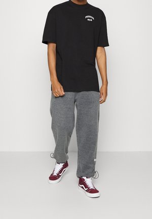 Camiseta de algodón negra con estampado gráfico blanco en el pecho, combinada con pantalones de chándal grises y zapatillas burdeos con acentos y cordones blancos.