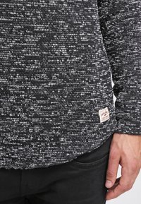 Detailní záběr na ruku opřenou poblíž kapsy černých kalhot, oblečenou v texturovaném černobílém pleteném svetru se malou látkovou značkovou etiketou.