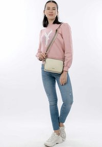 Felpa rosa chiaro con dettagli ricamati, jeans fitted blu medio e sneakers bianche. Borsa a tracolla crema con bordi neri.