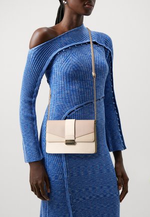 Femme portant une robe bleue côtelée, épaules dénudées, et un petit sac bandoulière beige avec une fermeture dorée et une chaîne strap.