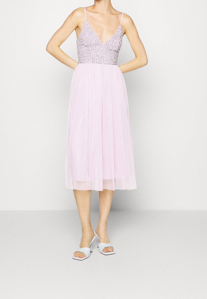 Mujer luciendo un vestido sin mangas de color lavanda con un corpete adornado y una falda midi transparente, acompañada de sandalias de tacón azul claro.
