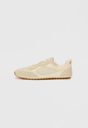 Beige athletic shoes with suede and mesh materials, rounded toe shape, lace-up design, and a brown rubber sole. Features subtle logo detail.