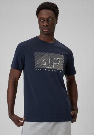 Mężczyzna ubrany w granatowy t-shirt z białym geometrycznym logo "4F" i napisem "Sportswear Est. 1995", w połączeniu z jasnoszarymi szortami.