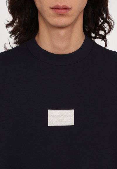 T-shirt en coton marine avec un col rond, présentant une étiquette texturée blanche avec le nom de la marque positionné au centre de la poitrine.