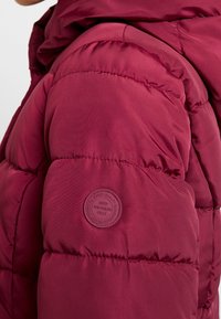 Burgunderfarbige gepolsterte Jacke mit einem runden Aufnäher am Ärmel, der mit geprägtem Text versehen ist. Glatte Textur mit gesteppten Abschnitten zur Isolierung.