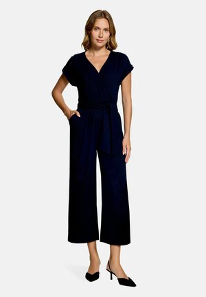 Kvinde iført en sort kortærmet slå-om buksedragt med vide ben og sorte slingback hæle, stående med den ene hånd i lommen.