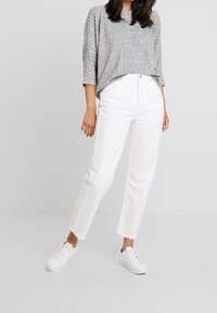 Maglietta grigia a maniche lunghe con texture, jeans bianchi a vita alta con orli sfrangiati e sneakers bianche. Design minimalista e colori neutri.