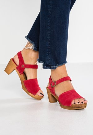Czerwone zamszowe sandały na obcasie z otwartym palcem i paskiem na kostce, z drewnianym obcasem i zapięciem na klamrę. Noszone z postrzępionymi dżinsami.