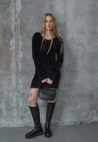 Black fuzzy dress with long sleeves and a fringe hem, paired with knee-high black leather boots and a small black handbag.
