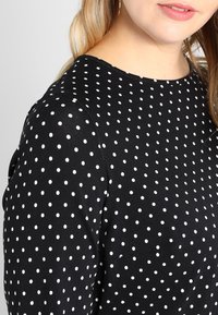 Černý top z látky s bílými puntíky, kulatým výstřihem a krátkými rukávy. Hladká textura, neformální design s volným střihem.