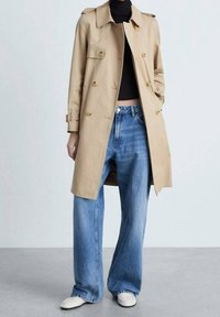 Manteau beige à double boutonnage, avec un col et des épaulettes, porté sur un haut noir et un jean bleu à jambes larges, associé à des chaussures blanches.