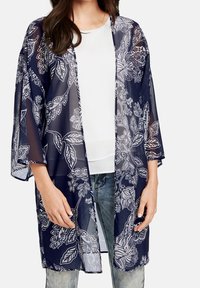 Kobieta w białej bluzce i wyblakłych niebieskich dżinsach, nosząca granatowy, przejrzysty kardigan w kwiaty w stylu kimono.