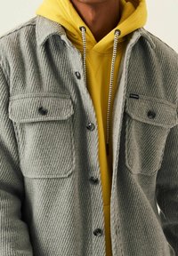 Gray houndstooth shirt jacket with two chest pockets, black buttons, and soft texture layered over a bright yellow hoodie with drawstrings.