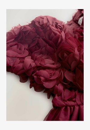 Burgundijas krāsu kleita ar slāņotām auduma ziedu detaļām, kas izceļas ar caurspīdīgu tekstūru un viļņotu apakšmali. Ziedu akcenti rada trīsdimensionālu efektu.