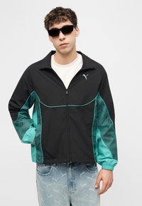 Jeune homme portant une veste zippée Puma noire et turquoise, un t-shirt blanc, un jean à motifs et des lunettes de soleil noires, debout avec une main dans la poche.