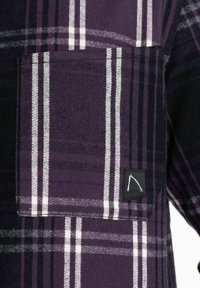 Lila und schwarze Karobluse mit einer Tasche, die weiße Streifen aufweist, und einem kleinen schwarzen Patch mit einem minimalistischen weißen Logo.