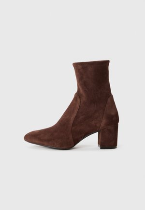 Bota de tobillo de ante marrón con punta afilada, lados elásticos y tacón cuadrado. La parte superior tiene una textura suave y una silueta ajustada.
