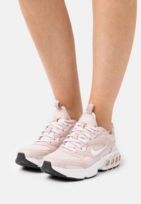 Scarpe Nike rosa chiaro con superiore in rete, suole bianche spesse e dettagli in gomma. Presentano un logo lateralmente e lacci spessi per supporto.