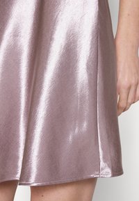 Skinande mauve satängkjol med en synlig söm, visad från mitt lår till midjan bredvid en hand och en bakgrund av blek hud.