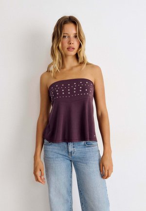 Junge Frau trägt ein ärmelloses lila Top mit silbernen Nieten und hellblaue Jeans und steht vor einem schlichten weißen Hintergrund.