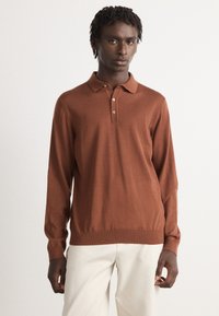 Junger Mann in einem rostbraunen Langarm-Polohemd und creamfarbenen Hosen, der vor einem schlichten hellen Hintergrund steht.