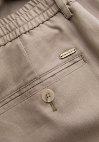 Pantalon en tissu beige avec ceinture élastique, poche arrière avec bouton et petite étiquette métallique portant la mention "QUINTESSENCE."