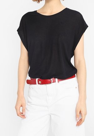 Femme portant un haut noir à manches courtes, ample, rentré dans un pantalon blanc taille haute avec une ceinture rouge.