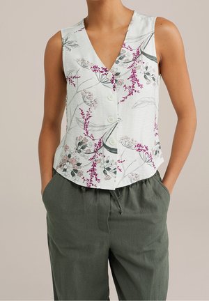 Blouse blanche sans manches avec imprimé floral rose et gris et boutons, portée avec un pantalon vert foncé à cordon, par une personne avec les mains dans les poches.