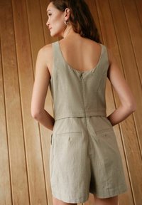 Ljus beige ärmlös jumpsuit med djup rygg, texturerad tyg och korta byxor. Har en dragkedja i ryggen och sidofickor.