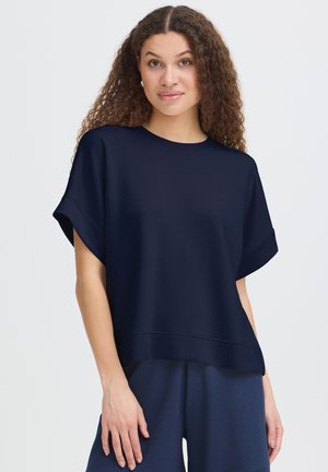 Frau mit lockigem Haar, die ein lockeres dunkelblaues Kurzarmoberteil und passende dunkelblaue Hosen trägt, steht vor einem einfarbigen Hintergrund.