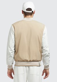 Jachetă bomber bej și albă cu manșete și tiv din material ribbed, având o textură netedă și mâneci contrastante. Guler ajustat și detaliu de branding.