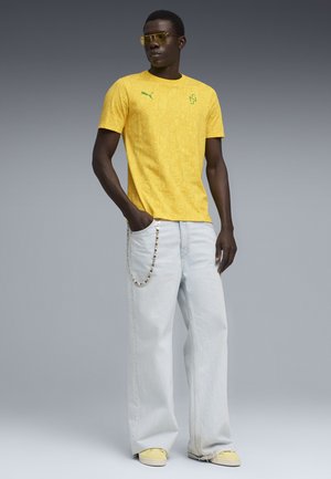 Homme portant un t-shirt Puma jaune à motif, un jean large bleu clair avec un accessoire chaîne, des baskets jaunes et des lunettes de soleil teintées jaunes, debout avec une main dans la poche.
