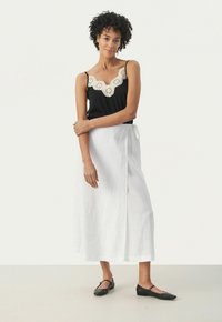 Black tank top with a scalloped, lace-like neckline, paired with a white, knee-length wrap skirt. Black flat shoes complete the look.