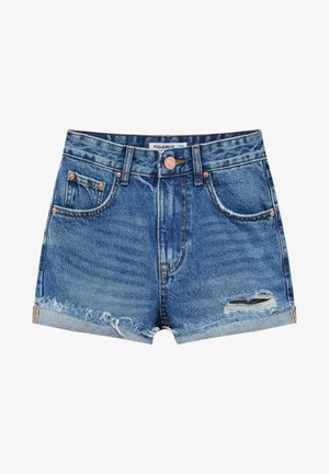 Woman standing with arms crossed wearing a white cropped t-shirt, denim cut-off shorts, white socks, and white sneakers.