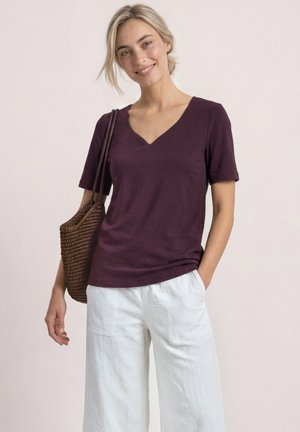 Jeune femme souriante portant un t-shirt violet à col en V, un pantalon blanc à jambes larges, portant un sac à bandoulière tressé marron sur fond clair.